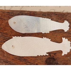 2 fichas decorativas de nácar con forma de pez, adornadas con pájaros y escamas.