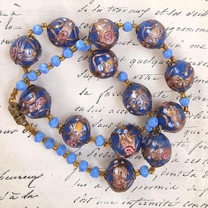 May include: A vintage necklace featuring large, round, cobalt blue beads with intricate gold and pink floral designs. Smaller light blue beads and gold-toned spacers connect the larger beads. The necklace is secured with a gold-toned clasp.