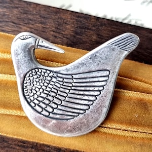 Könnte beinhalten: Silberfarbene Vogelbrosche. Die Brosche zeigt einen Vogel mit langem Schnabel, Auge und Flügeldarstellung. Die Brosche liegt auf einer goldfarbenen Samtoberfläche.