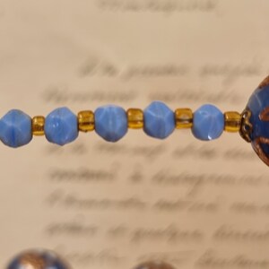 May include: Close-up of a necklace featuring light blue faceted beads and gold-colored spacers. The necklace has a vintage aesthetic, with a blurred background of handwritten text.