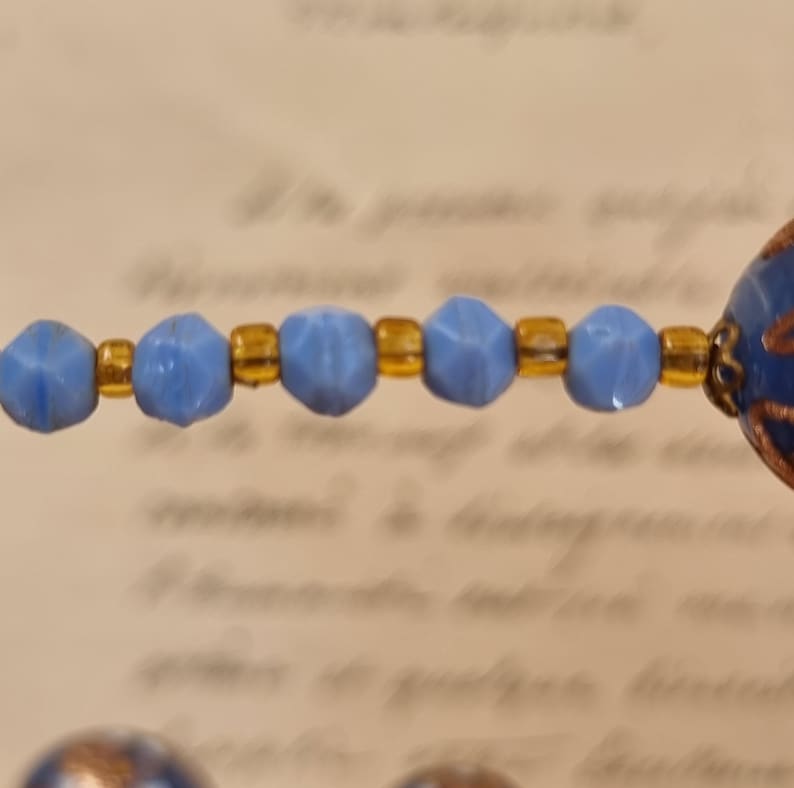 May include: Close-up of a necklace featuring light blue faceted beads and gold-colored spacers. The necklace has a vintage aesthetic, with a blurred background of handwritten text.