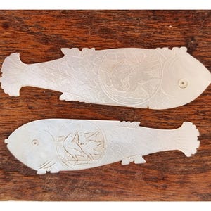 May include: Two white, fish-shaped decorative pieces with intricate carved details. Each fish features a circular design with a bird motif. The pieces are set against a dark wood background, highlighting the delicate carvings and shapes.