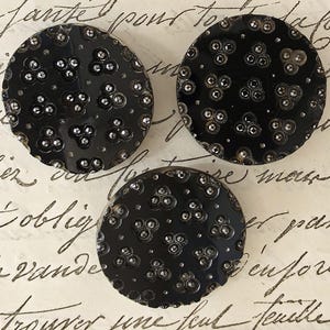 May include: Three black, circular buttons with a textured surface and a pattern of small, silver embellishments. The buttons are on a background with handwritten script.