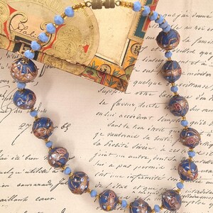 May include: A vintage necklace featuring dark blue, round beads with gold and red accents. The necklace has smaller light blue beads between the larger ones and a gold clasp. The background includes a vintage tobacco box and handwritten script.