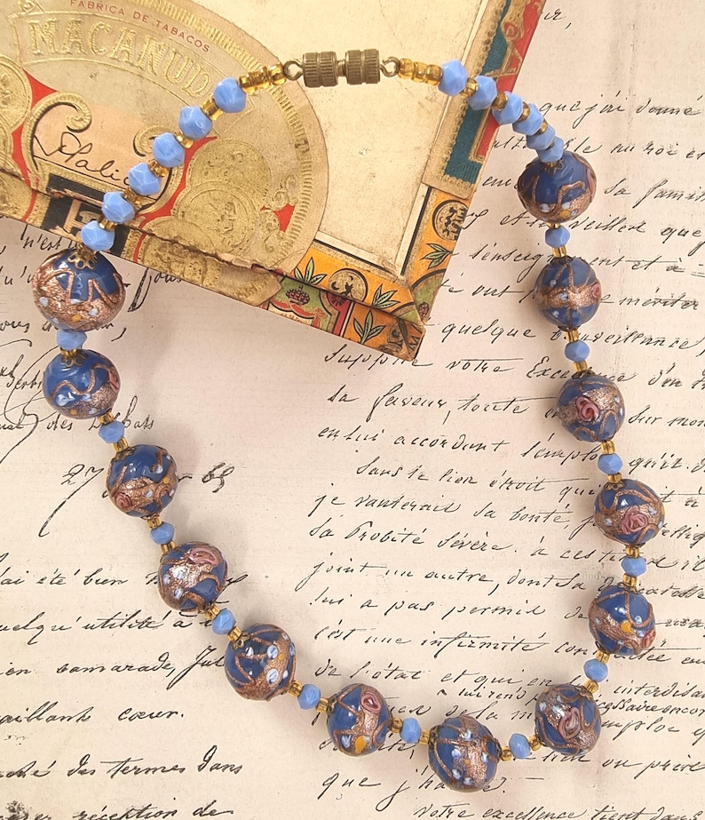 May include: A vintage necklace featuring dark blue, round beads with gold and red accents. The necklace has smaller light blue beads between the larger ones and a gold clasp. The background includes a vintage tobacco box and handwritten script.
