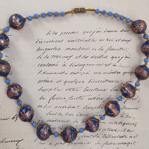 May include: A vintage necklace featuring large, dark blue beads with intricate gold and red designs. Smaller light blue beads and gold-toned spacers connect the larger beads. The necklace is displayed on a handwritten document.
