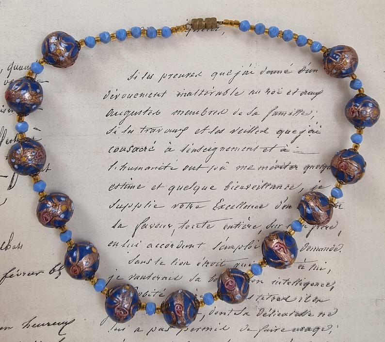 May include: A vintage necklace featuring large, dark blue beads with intricate gold and red designs. Smaller light blue beads and gold-toned spacers connect the larger beads. The necklace is displayed on a handwritten document.