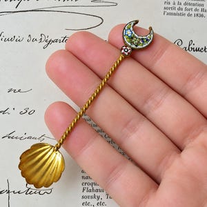 Può includere: Un cucchiaio in metallo dorato con manico intrecciato, coppa a forma di conchiglia e un terminale a mezzaluna decorato con motivi floreali in micro-mosaico colorato e un piccolo intarsio circolare a forma di stella.