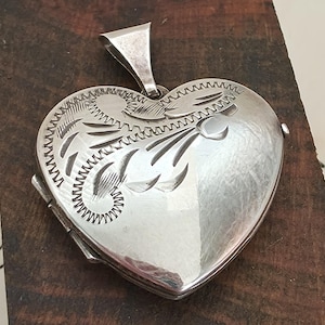 May include: A heart-shaped silver locket pendant featuring an engraved floral pattern on the front surface. The locket includes a triangular bail at the top for attachment to a chain, with visible hinge details along the left side of the metal frame.