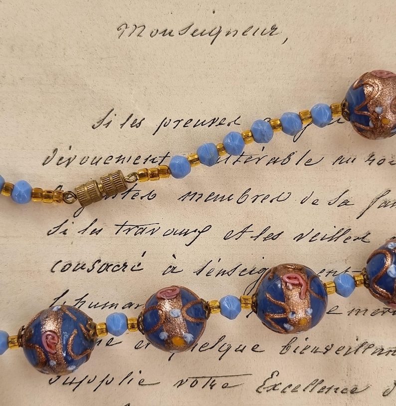 May include: A vintage necklace featuring blue and gold beads with decorative, patterned blue and copper-colored focal beads. The necklace is displayed on a handwritten document with French text.