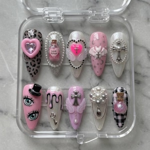 May include: A set of ten press-on nails in a clear plastic case. Designs include a leopard print, a pink heart, a love potion bottle, a heart with wings, a cross, a top hat, dripping paint, a key, a cross with wings, and a black and white checkered pattern.