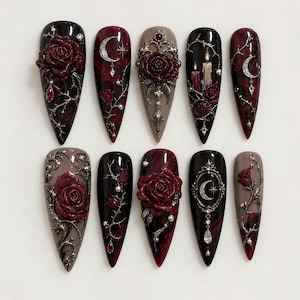May include: A set of ten stiletto-shaped press-on nails featuring dark red and black marble patterns, adorned with three-dimensional silver metallic rose embellishments, crescent moon charms, lit candle motifs, and small clear crystal accents.