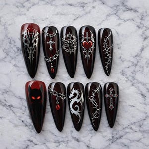 May include: A set of ten long, pointed, dark red artificial nails. Each nail features a unique design in silver or white, including chains, crosses, a pentagram, a dragon, and a devil face. The nails are arranged on a marble surface.