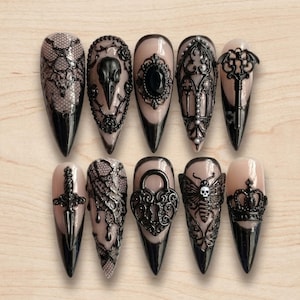 May include: A set of ten stiletto-shaped press-on nails featuring a nude base with intricate black 3D embellishments, including a bird skull, ornate frames, a key, a dagger, a heart-shaped padlock, a moth with a skull, and a crown.