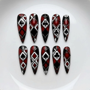 May include: Ten long, almond-shaped artificial nails. Each nail features a design with red, black, and white playing card suit symbols, including hearts, diamonds, spades, and clubs. The nails have a glossy finish and a dark border.