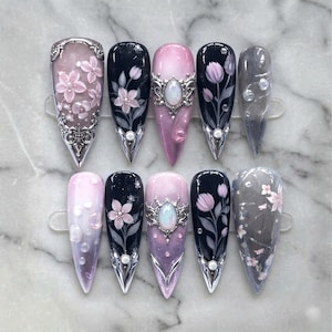 May include: A set of ten long, pointed artificial nails. Designs include pink and black floral patterns, silver embellishments, and gradient effects. Some nails feature 3D floral accents and pearlescent details.