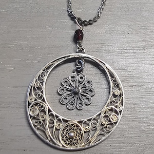 Op de afbeelding: Zilveren filigraanhangerketting met een cirkelvormig ontwerp. De hanger heeft een grote opengewerkte cirkel met ingewikkelde details, een kleinere bloemvormige bedel en een donkerrode edelsteen. De ketting is ook zilver.