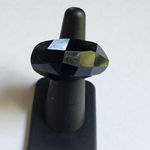 May include: A black ring featuring a faceted, geometric surface design. The ring is constructed with multiple flat, angled planes that create a reflective, multi-sided appearance. The band is solid black and displays a polished, dark finish throughout.