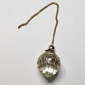 Könnte beinhalten: Eine goldfarbene Metallkette, die an einer kugelförmigen, facettierten, klaren Glasperle befestigt ist. Die Perle ist von einem feinen goldfarbenen Drahtnetz umhüllt und wird oben von einer kleinen, glatten silberfarbenen Metallkappe gehalten.
