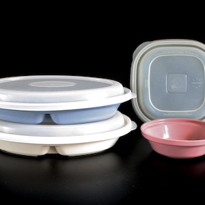 May include: Three plastic food containers with lids. One is a round container with a divided base and a white lid. Another is a round container with a blue lid. The third is a square container with a gray lid. The containers are on a black surface.