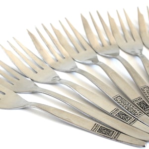 May include: A set of stainless steel forks with a decorative pattern on the handles. The forks are arranged diagonally, with the tines pointing upwards. A spoon is visible in the background. The cutlery is set against a white background.
