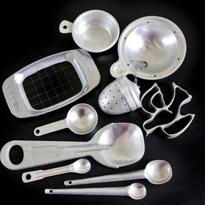 May include: A set of eight silver-colored kitchen utensils, including a measuring spoon, a tea infuser, a small bowl, a cookie cutter, and a strainer. The utensils are arranged on a black surface.