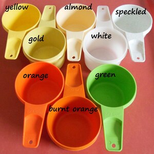 May include: Set of 8 vintage measuring cups in various colors, including yellow, gold, almond, white, speckled, orange, burnt orange, and green. Each cup has a handle and a measurement marking.