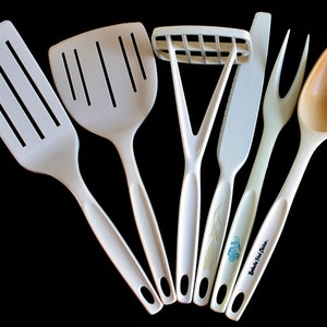 Puede incluir: Un juego de seis utensilios de cocina blancos, que incluyen una espátula ranurada, una espátula sólida, un machacador de patatas, un cuchillo, un tenedor y una cuchara de madera. Los utensilios son todos blancos con mangos negros y presentan un diseño floral azul y blanco.