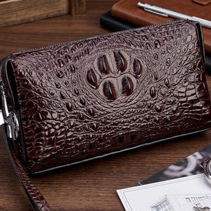 May include: A brown leather clutch with a crocodile skin pattern. The clutch has a zipper closure and a wrist strap. The front of the clutch features a raised, textured design. The overall design is a dark brown color.