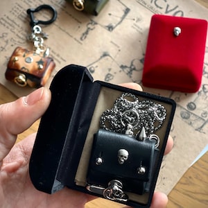 May include: A black leather miniature purse with silver skull accents, displayed in a black velvet box. Other miniature purses in brown, green, and red are also visible, along with a keychain. The word "SACVO" is visible on the background.