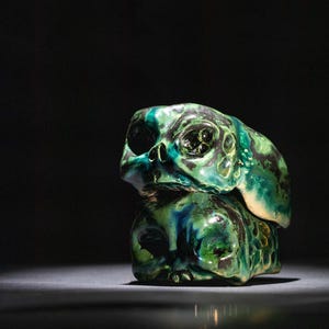 May include: Two ceramic skull sculptures stacked on top of each other. The skulls are glazed in shades of green and blue, with a glossy finish. The sculpture has a unique, artistic design.