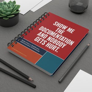 May include: A spiral-bound notebook with a red cover featuring the white text: SHOW ME THE DOCUMENTATION AND NOBODY GETS HURT. Below this, a dark blue section reads: Notes From a Healthcare Administrative Professional, with CODEMEDDIGITAL printed.