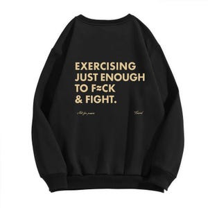 May include: Black sweatshirt with the text "EXERCISING JUST ENOUGH TO F≈CK & FIGHT" in a gold color. Below the text are the words "Not for panics" and "Friend".
