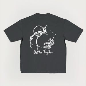 Puede incluir: Camiseta gris oscuro con un dise&ntilde;o blanco de un esqueleto abrazando otra forma, con el texto "Better Together". El dise&ntilde;o est&aacute; centrado en la parte trasera de la camiseta, creando un impacto visual audaz.