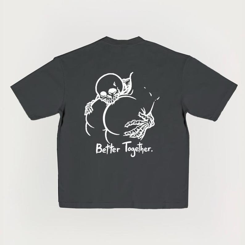 Puede incluir: Camiseta gris oscuro con un dise&ntilde;o blanco de un esqueleto abrazando otra forma, con el texto "Better Together". El dise&ntilde;o est&aacute; centrado en la parte trasera de la camiseta, creando un impacto visual audaz.