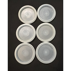 May include: Six clear, round Tupperware lids are arranged on a dark surface. Each lid has a white rim and a slightly textured center. The text "Tupperware" and "Made in U.S.A." are visible on the lids.