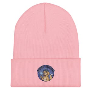 Puede incluir: Un gorro rosa claro con un borde doblado. El gorro presenta un parche bordado con las palabras "SNIFF SQUAD" alrededor de un perro de dibujos animados que sostiene una lupa. El perro lleva un sombrero de detective.