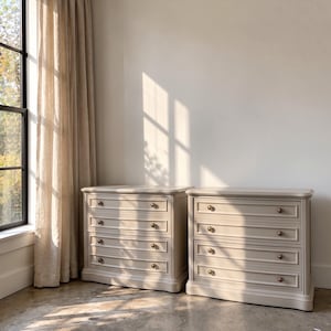 May include: Two matching light beige wooden chests of drawers, each featuring four horizontal drawers with recessed panel detailing and small, round, metallic knobs. The furniture pieces have a smooth, matte finish and a rectangular shape.