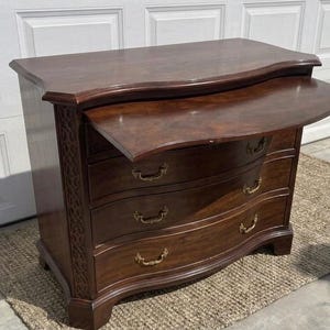 May include: A dark brown wooden chest of drawers featuring a serpentine front, three lower drawers with brass-toned metal pulls, and a pull-out writing surface at the top. The side panels display an intricate carved lattice pattern design.