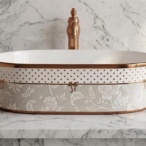 May include: An oval ceramic vessel sink featuring a white interior and a copper-toned rim. The exterior displays a white band with black polka dots above a light gray section decorated with a white floral pattern and small copper-colored bows.