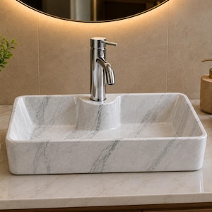 May include: A rectangular bathroom sink with a white and gray marble pattern. The basin features a central raised platform that holds a polished chrome faucet. The sink has a smooth, glossy finish and a wide, flat rim, presenting a clean design.