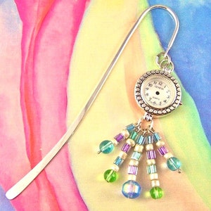 May include: Silver bookmark with a small watch charm and a dangling cluster of colorful beads. The watch face is white with black numbers and hands. The beads are green, blue, purple, and white.