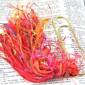 May include: A close-up of a book with a colorful bookmark made of red, orange, pink, and yellow yarn. The bookmark is held in place by a gold paper clip. The text on the page reads "For the multitude of thy enchantments."