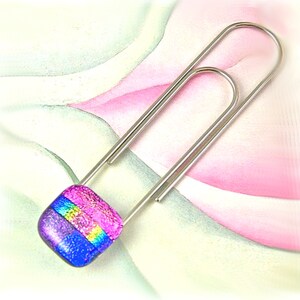May include: A silver paperclip with a square piece of iridescent glass attached to it. The glass is pink, blue, and yellow.