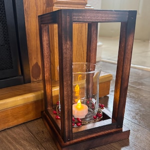 May include: A dark brown wooden lantern with a glass cylinder inside, holding a lit candle. The candle sits on a bed of red berries. The lantern is rectangular and sits on a wooden floor.
