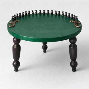 May include: A small, round table with a dark green tabletop and dark brown legs. The tabletop is edged with small, dark pegs and decorative brass accents. The table has four turned legs, each with a bulbous design. The table is a decorative furniture piece.