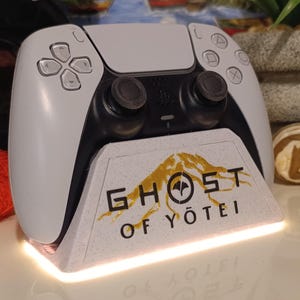 Peut inclure: Manette blanche et noire PlayStation 5 sur une base blanche avec le texte "GHOST OF YŌTEI" en noir et or. La base a une lumière brillante en bas. Accessoire de jeu.