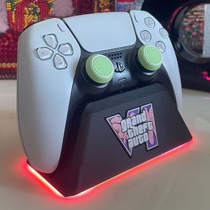 Peut inclure: Une manette de jeu blanche et noire est placée sur une station de charge noire arborant le logo Grand Theft Auto V. La station émet une lumière rouge à sa base et la manette est équipée de capuchons de joystick texturés vert clair.
