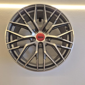 Könnte beinhalten: Eine silber-schwarze Leichtmetallfelge mit Mehrspeichen-Design. Die Radkappe ist rot mit den weißen Buchstaben "MAM". Das Rad ist an einer weißen Wand befestigt.