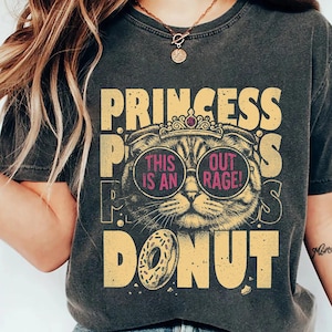 May include: Dark gray t-shirt with a graphic design featuring a cat wearing sunglasses and a crown. The text reads "PRINCESS, THIS IS AN OUT RAGE! DONUT". The design is in shades of yellow and pink.